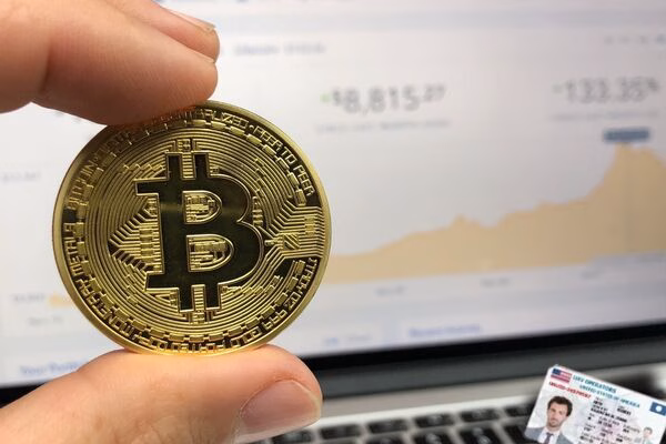 A person holds Bitcoin while viewing a laptop screen, with an ID visible.
