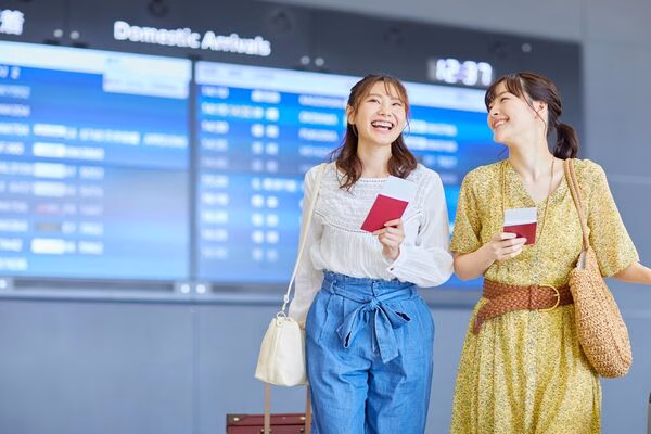 Two women smiling at airport, holding passports and plane tickets.
