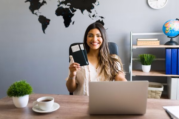 Woman smiling at desk while holding plane tickets confidently.