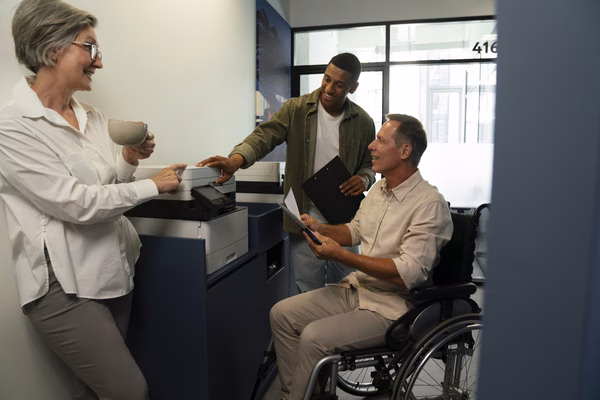 Man in wheelchair discussing plane tickets with coworkers at office.

