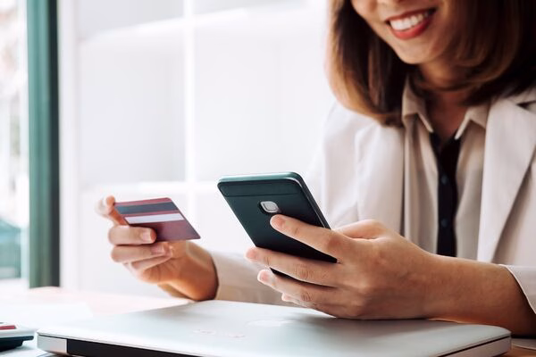 Mujer sonriente que usa un teléfono y una tarjeta para pagos transfronterizos.
