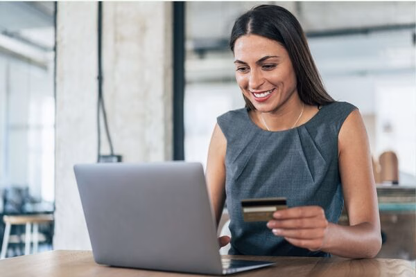 Una mujer sonriente realiza pagos transfronterizos usando una computadora portátil y una tarjeta.
