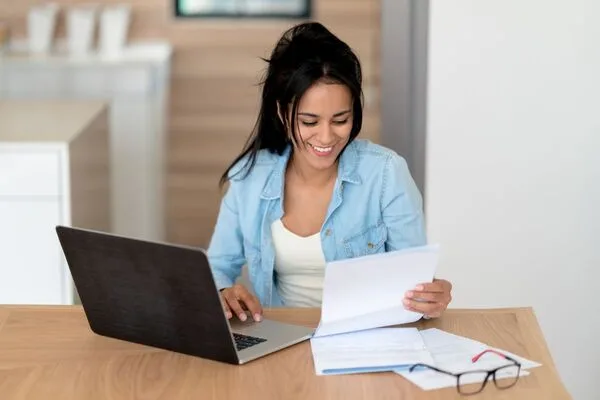 Smiling woman reviewing Money Order documents while using laptop.